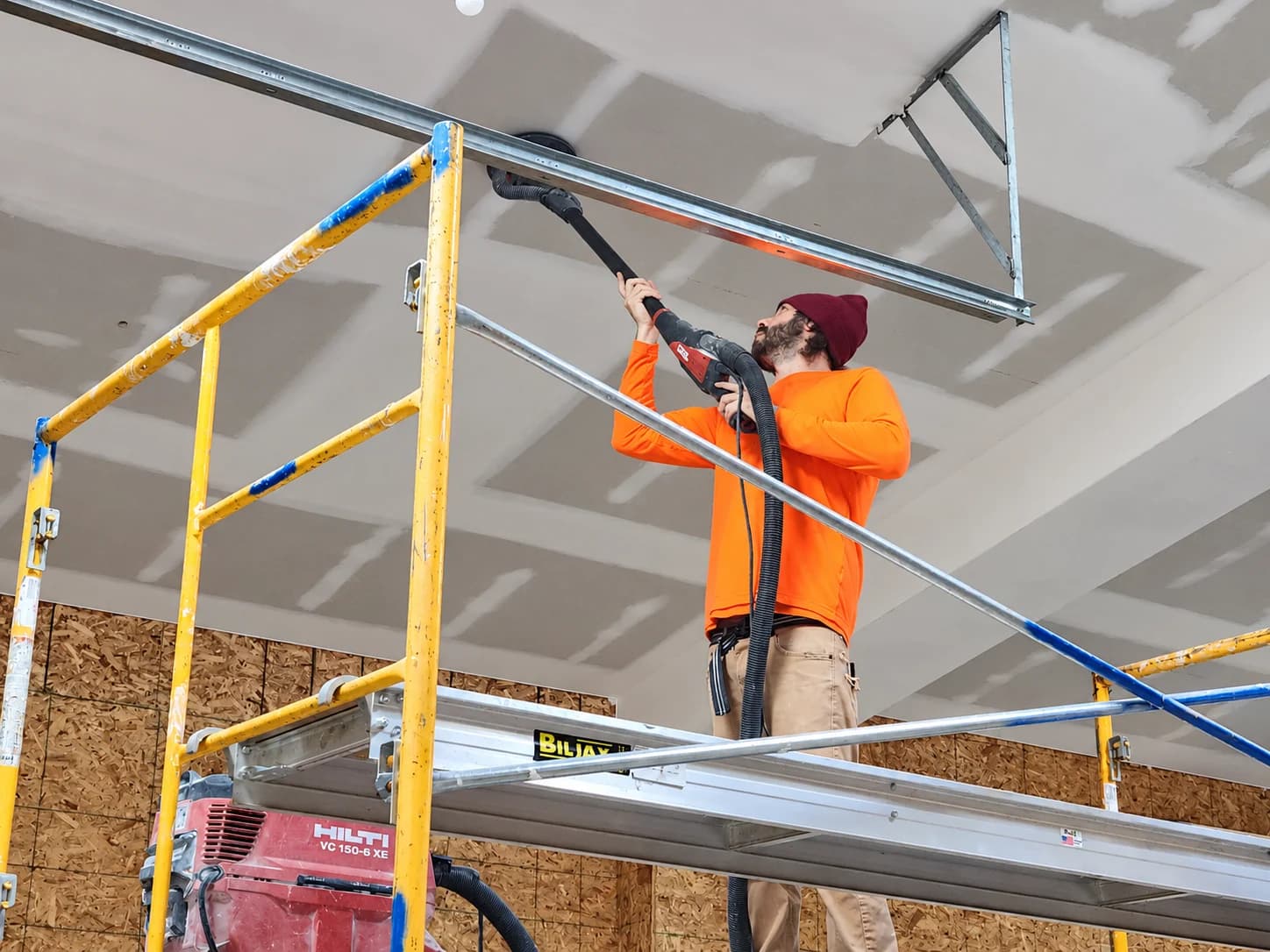 A worker sanding drywall seams on a ceiling from a scaffold